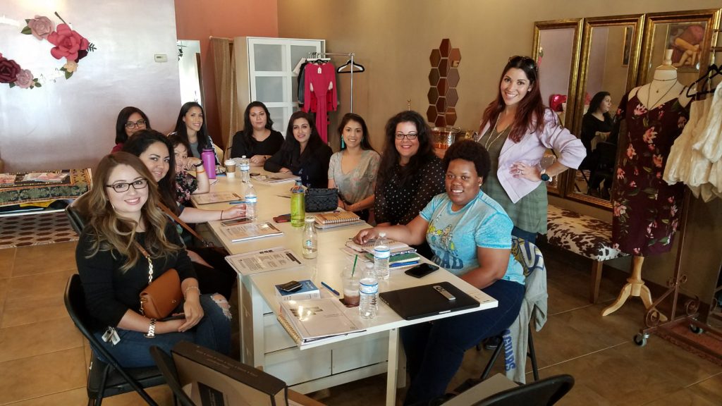 A group of beautiful, smiling, diverse women sit at a table, ready to start a blog class.