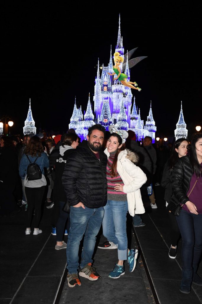 Experience Gifts for Valentine's Day idea: A beautiful smiling couple pose for a photo in front of Cinderella's Castle at Disney's Magic Kingdom. It is lit up with twinkling white lights during winter.